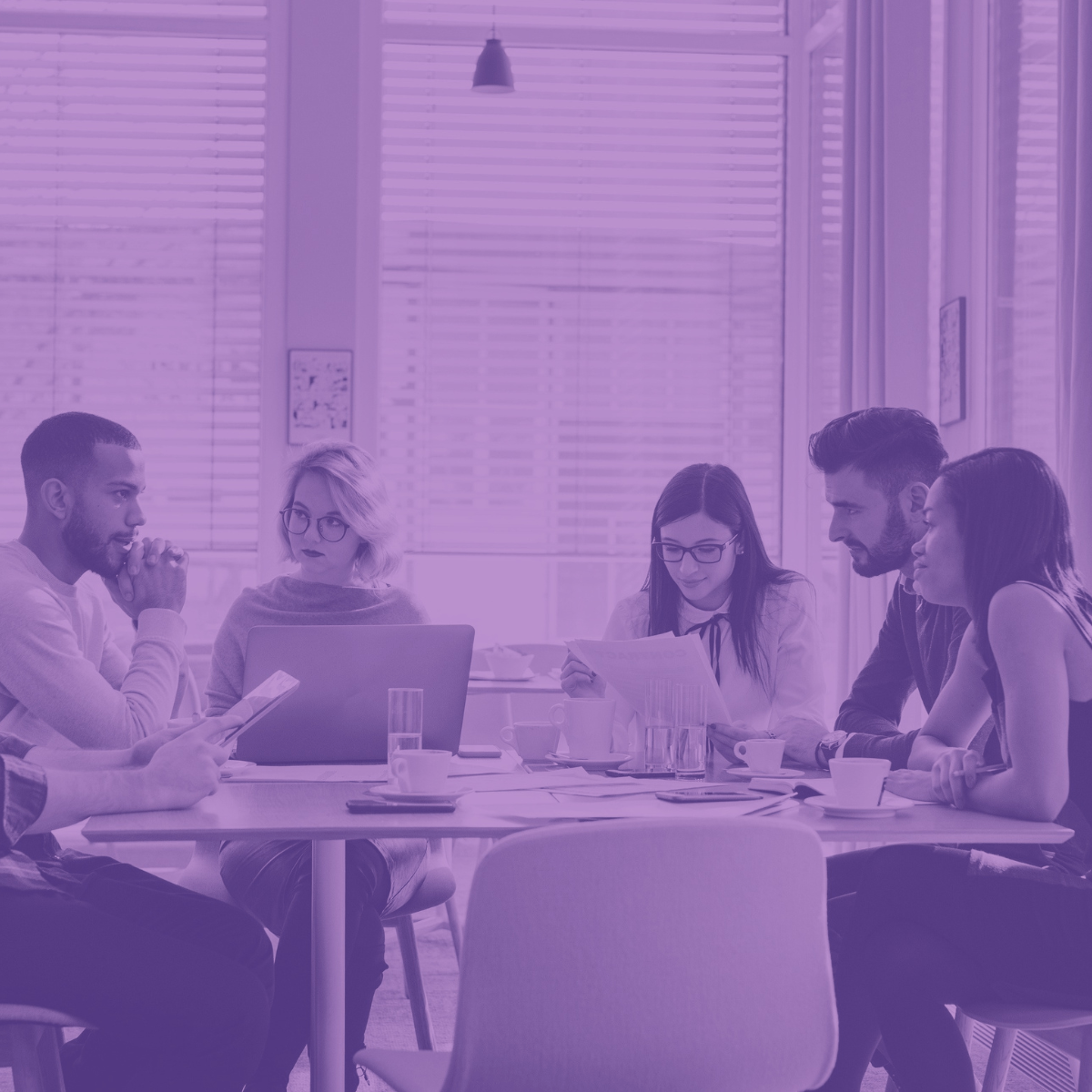 A group of five people are sitting around a table working and talking intently with a purple filter overlay